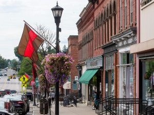A small-town street lined with historic brick buildings, featuring shops with green awnings and large windows. A lamppost adorned with hanging flower baskets and a red and brown flag adds charm to the scene.