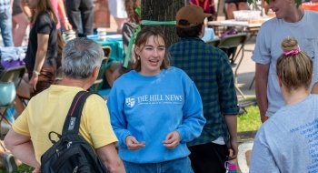 A student person wearing a blue sweatshirt with 'The Hill News' logo is engaging in conversation at an outdoor event. They are surrounded by other people, some seated and some standing, with tables and booths in the background. The atmosphere is casual and lively.