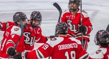 A group of ice hockey players in red and white jerseys with 'SLU' on the front celebrate on the ice. One player, wearing a 'C' on their jersey, indicating they are the captain, is smiling widely. The players wear black helmets and gloves, and the background shows the ice rink surface.