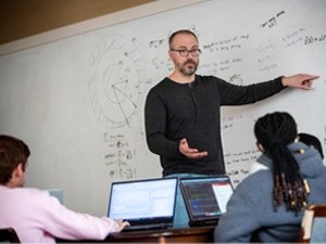A statistics professor, wearing glasses and a dark grey henley t-shirt, stands in front of his class and points to calculations on the white board.
