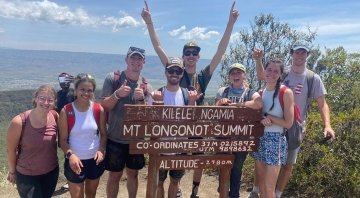 A group of Saint Lawrence students stand in front of a sign at the summit of a mountain in Kenya. A person in the background takes a photo while the group smiles and raises their arms in the air.