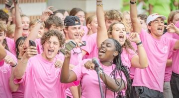 A large group of Saint Lawrence University students wearing matching pink shirts cheer and smile during an outdoor event, with one person singing into a microphone and others holding up phones.