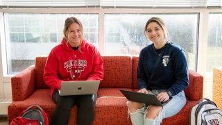 Two students sit on a red couch in a bright, airy study space with lots of windows. They both type on laptops. 