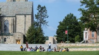 A professor teaches a class outdoors on a beautiful late summer day on the quad, framed by a stone chapel and brick academic building.