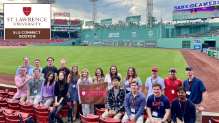 A group of 20 students holding a St. Lawrence flag sits at the edge of the field at Fenway Park, with the iconic "Green Monster" in left field visible in the background.