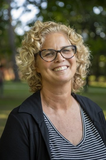 Headshot of Professor Liz Regosin, standing against a backdrop of trees. She has curly hair, glasses, and wears a black and white striped shirt with a black blouse.