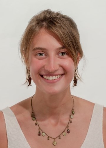 Headshot of Ellie Shaw, with her hair pulled back, a simple white tank top, and a tear-drop necklace with green gemstones.