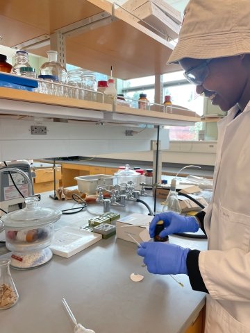 Leah Biwot, wearing a tan bucket hat and white lab coat, works in a lab next to chemistry equipment.