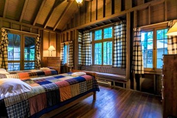 The interior of a lodge cabin. Two single beds with blue and red plaid quilts. Navy blue plaid curtains hang from four large windows.