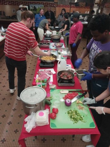 Students gather around a red table, chopping vegetables and cooking tasty food.