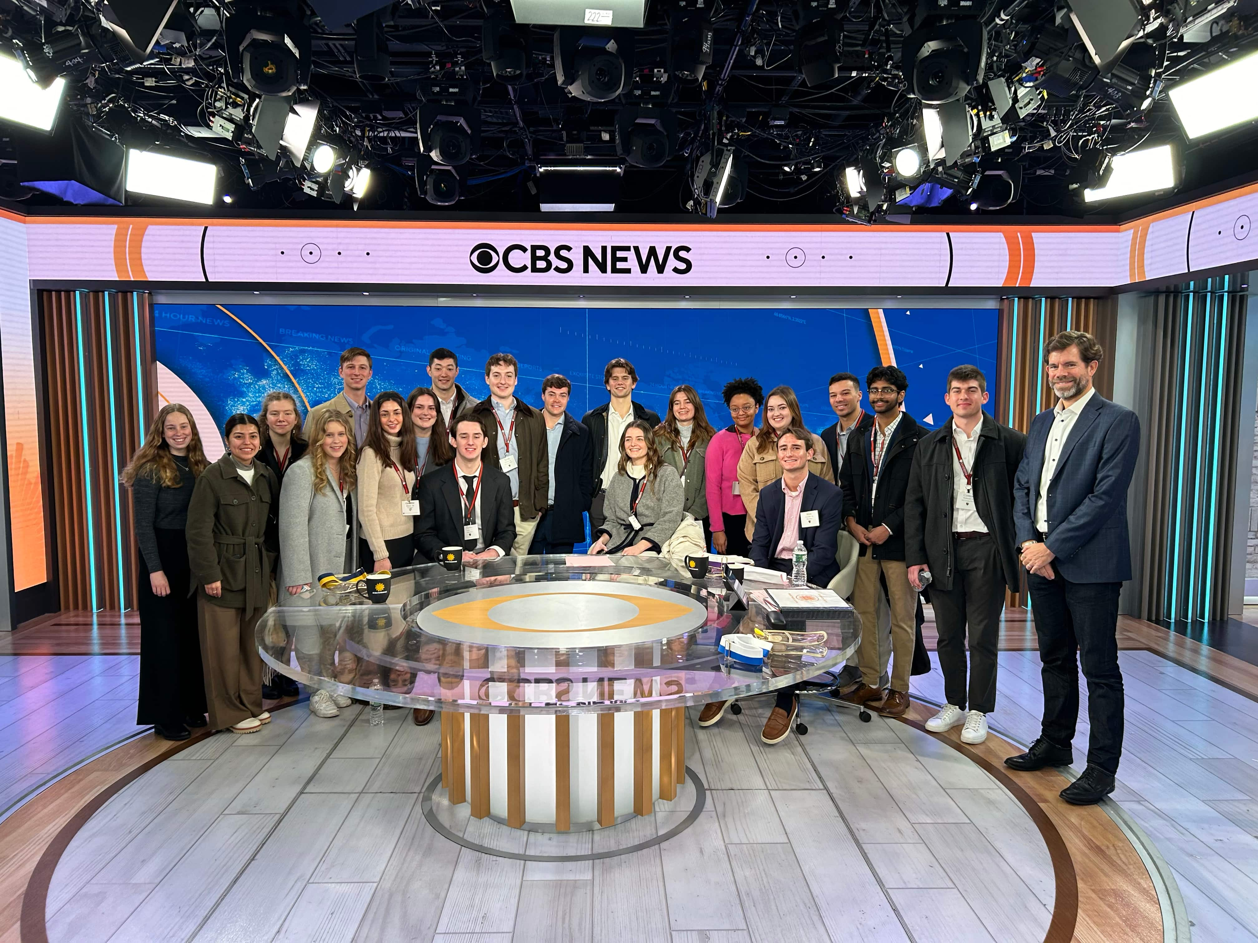 A group of people stands in a semi-circle around a large, round news desk inside the CBS News studio. The group consists of men and women of varying ages, all dressed in business casual attire. The studio is brightly lit with professional lighting equipment, and the CBS News logo is prominently displayed on a large screen in the background.