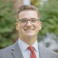 Headshot of Jaron Belmore, a young man with light brown hair, wearing glasses, a gray suit, a white shirt, and a red tie with small emblems. He is smiling and standing in front of a blurred, outdoor background with greenery.