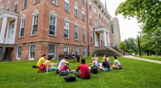 A professor sits on a bench outside of Richardson Hall, delivering a lecture to a group of students seated around her. It's a warm late summer day, perfect for an outdoor class.