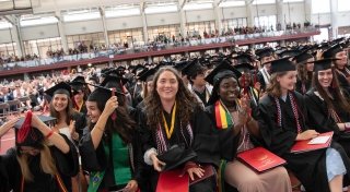 A group of Saint Lawrence students, wearing graduation regalia, sit in the front row at Commencement smiling as they flip their tassles.