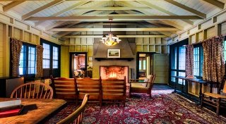 The living room of an Adirondack style cabin with wooden beams on the ceiling. A brick fire place has a warm, glowing fire while a several books rest on a table nearby.