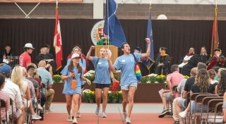 A group of energetic student leaders in light blue t-shirts and shorts march down an aisle, cheering and carrying a blue flag. The scene, from Matriculation 2023, takes place in a gymnasium, with faculty in regalia on stage and a "Home of the Saints" banner above. The audience watches the lively procession.