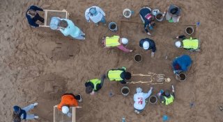 A group of people at an archaeological dig work around a partially uncovered skeleton in the sand. Some wear safety vests and use tools to brush away sand, while others sift dirt through a wooden frame. The scene is viewed from above.