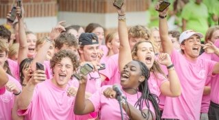 A group of Saint Lawrence University students wearing pink shirts celebrates together, many holding up their phones and cheering enthusiastically. The scene is energetic, with smiling faces and raised arms, creating a lively and joyful atmosphere. One student stands in front of a microphone, adding to the sense of excitement.