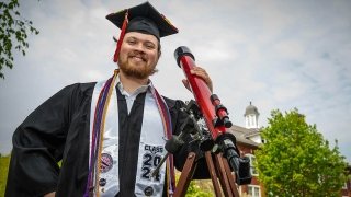 Tyler Karaskinski, dressed in full graduation regalia, stands next to a red telescope in front of a brick building. It's a cloudy day with some sun peeking through.