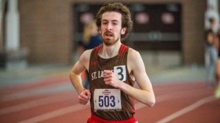  A young male athlete wearing a brown and red St. Lawrence University track uniform with the number 503, running on an indoor track with a determined expression. The background is blurred, highlighting his focus and motion.