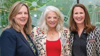 President Morris stands on the left, smiling warmly, wearing a dark blazer and a patterned blouse. In the center is Sarah Johnson, wearing a floral-patterned jacket over a red top, with white hair styled loosely around her shoulders. On the right is Jennifer Curley Reichert, also smiling, wearing a patterned jacket over a black blouse. The three women are close together, appearing at a formal event, with a modern glass backdrop featuring geometric designs behind them. The atmosphere is professional yet frie