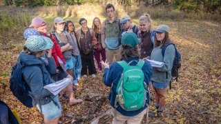 A group of students gathers in a circle outdoors, listening attentively to an instructor with a green backpack. The group is standing on a leaf-covered forest floor, holding notebooks and dressed for a field experience. Sunlight filters through the trees, creating a warm, autumn atmosphere.