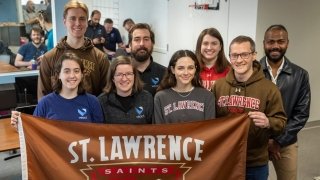 A group of Saint Lawrence University alums stand together and hold up a brown Saint Lawrence University flag. They are inside an office at a technology company.