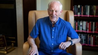 Thomas Wallace, wearing a blue button up t-shirt, sits in a leather chair in a library.