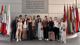 A group of individuals stands in front of the European Central Bank wall, with various national flags displayed on either side. They are smiling and dressed in a mix of casual and formal attire. The setting suggests a visit or event at the bank.