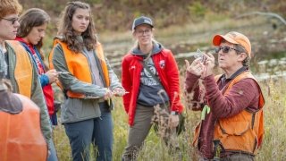 Erika Barthlemess, wearing a florescent orange vest, stands in front of a group of Saint Lawrence students and explains a research sample collection process. The group is outside in the woods.