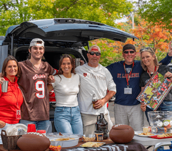 Saint Lawrence University students and parents tailgating together, posing in front of a car, with food and drinks on the table during Laurentian Weekend.