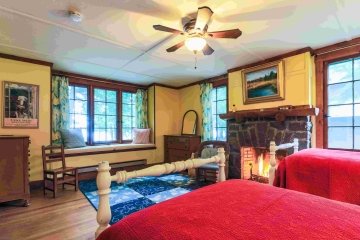 A yellow bedroom with a large window seat, a roaring brick fireplace, and two single beds with red quilts on them. 