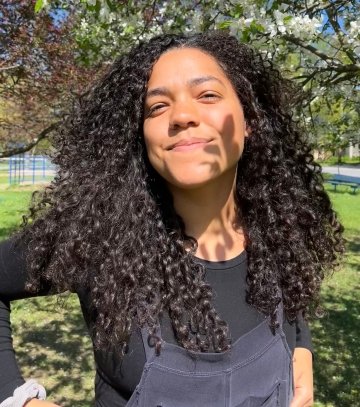 Adarra Smith, with long curly hair, stands under a flowering tree. She is wearing a black long-sleeved shirt under overalls.