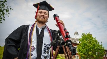 Tyler Karaskinski, dressed in full graduation regalia, stands next to a red telescope in front of a brick building. It's a cloudy day with some sun peeking through.