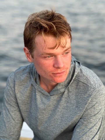 Headshot of Jax Lubkowitz, with dirty blonde hair and wearing a simply gray sweatshirt against a background of placid water.