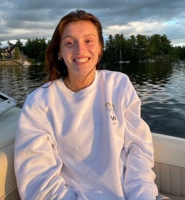 Alli Sibold, wearing an oversized white sweatshirt, sitting on a boat at dusk with a lake and trees behind her.