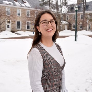 Headshot of Miso Wilson, wearing a white long-sleeve shirt and plaid dress. In the background, campus is covered in snow and there is a brick dorm building.