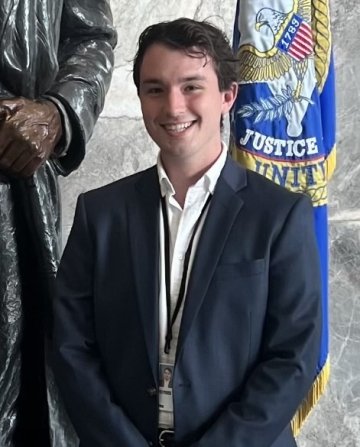 John Reece, where a suit jacket and white collared shirt, standing in front of a marble wall and a blue flag.