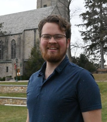 Headshot of Tyler Karasinski, wearing a navy blue polo and standing in front of a the stone chapel on the St. Lawrence campus.