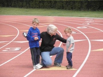A woman kneeling on a red running track, with her arms around her two young sons. Both boys are dressed casually in athletic clothing, and the scene takes place on a sunny day with a grassy area in the background.