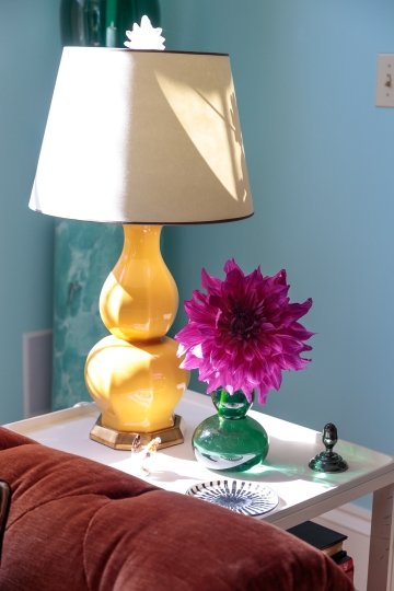 A brightly lit side table with a yellow lamp, featuring a beige lampshade, sits against a light blue wall. Next to the lamp is a green vase holding a large, vibrant purple flower. Additional items on the table include a small decorative plate, a dark metal figurine, and a few books partially visible below.