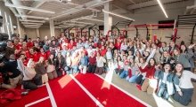 A large group of Saint Lawrence University students, many dressed in red and gray, gather excitedly in the campus fitness center, posing and cheering. They stand in front of workout equipment under bright overhead lights. The group is smiling and waving, creating a lively and celebratory atmosphere.