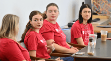 Four students sit at a table, engaged in a discussion. They are wearing matching red Saint Lawrence University shirts, and several iced coffee drinks are on the table. The background shows a bright classroom with a colorful abstract design on the wall.