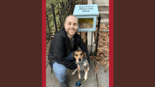 Sean Kelly crouches on a sidewalk, smiling while holding a dog on a leash. Next to them is a small blue box labeled "Stevie's Biscuit Bar," filled with dog treats. Fallen leaves and a black metal fence frame the scene.