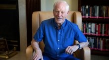 Thomas Wallace, wearing a blue button up t-shirt, sits in a leather chair in a library.