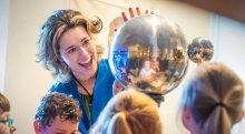 A smiling instructor in a blue jacket shows a group of children a reflective metal sphere during a science demonstration. The children look on curiously, and their reflections are visible in the shiny surface of the sphere.